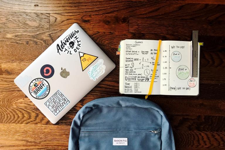 A laptop with stickers, a textbook with writing and diagrams and a blue backpack against a wood background
