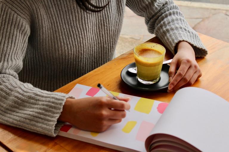 Student at desk with a coffee, writing on a pad with coloured blocks