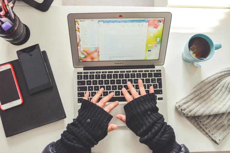Hands in fingerless gloves typing on a laptop with coffee and phone on desk
