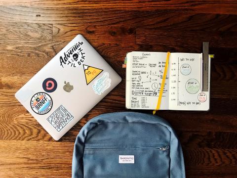 A laptop with stickers, a textbook with writing and diagrams and a blue backpack against a wood background